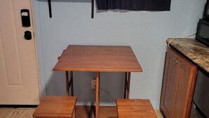 A compact kitchen with a wooden table and two stools, a window with curtains, and a door with a lock. - Central Maui Studio • Kitchenette + Queen Bed (Wailuku)