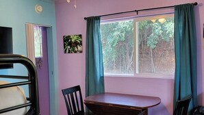 Ein Zimmer mit einem Etagenbett, einem Tisch mit zwei Stühlen, einem Deckenventilator, einem Fenster mit Vorhängen und einer kleinen Pflanze an der Wand.