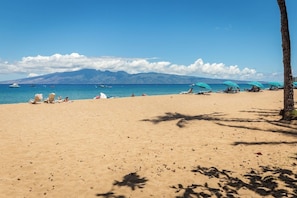 A beach with clear skies, a mountain range in the background, and several umbrellas and lounge chairs.
