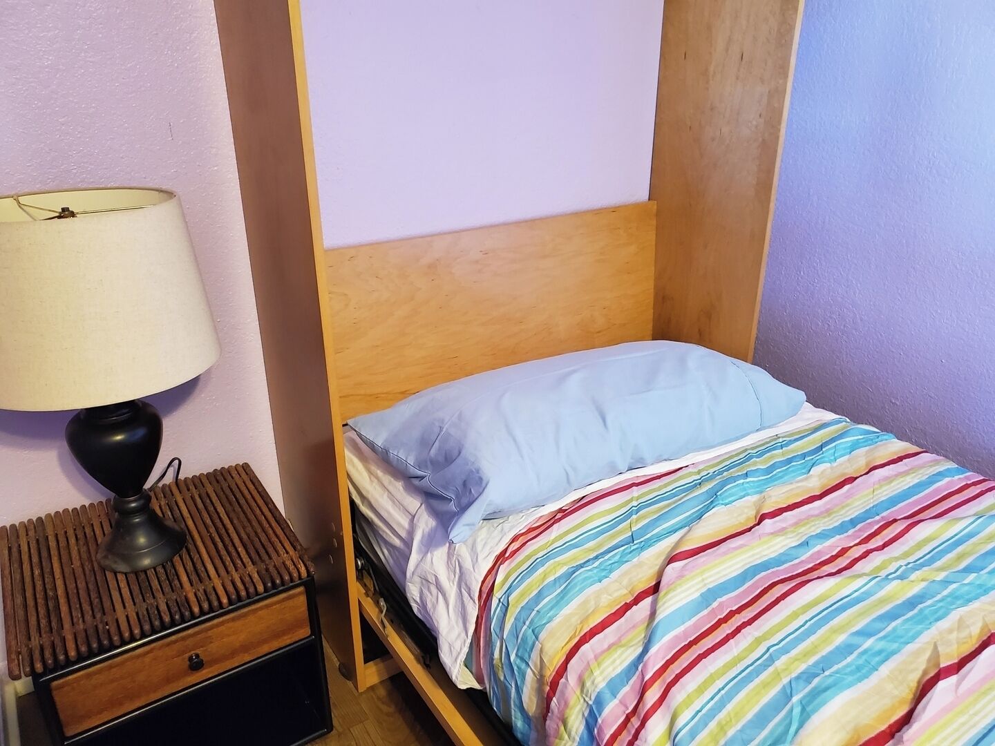 A single bed with a striped blanket, a bedside lamp, and a wooden headboard in a room with plain walls.