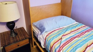 Un llit individual amb llençol estriat, una làmpada de nit i un panell de fusta al capçal en una habitació amb parets sense decoració.