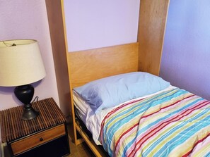 Una cama individual con una colcha a rayas, una lámpara de noche y un cabecero de madera en una habitación con paredes lisas.