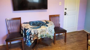 Una habitació amb televisió penjada a la paret, una porta i dues cadires amb un taulell cobert per un mantell estampat.