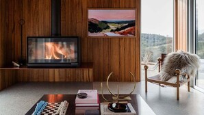 A modern living room with a fireplace, a painting, and a view through a large window. - Threefold - Freycinet Luxury Hosts (Swansea)