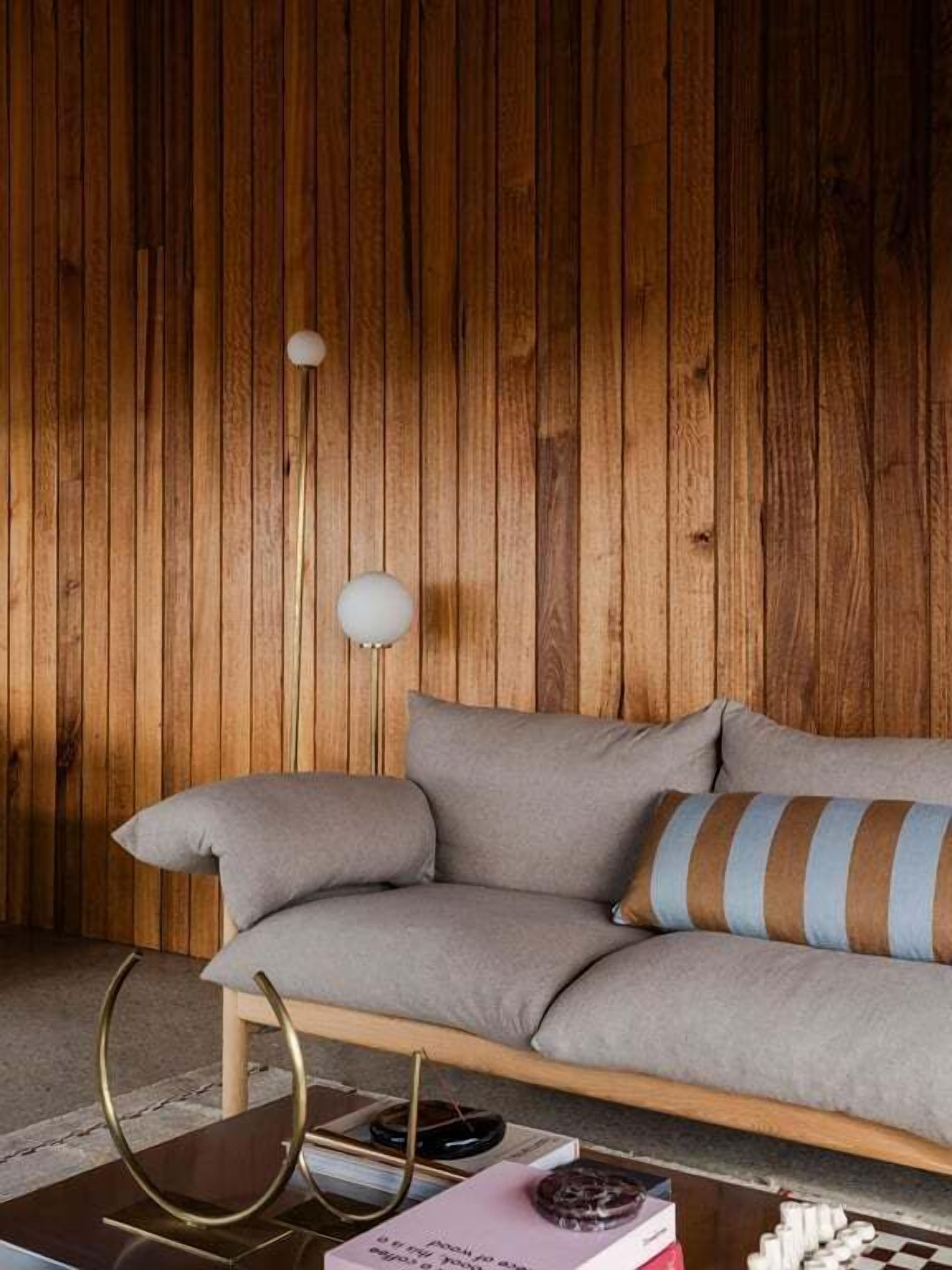 A room with a wooden paneled wall, a grey sofa with striped cushions, a round table with books and a globe, and a floor lamp.