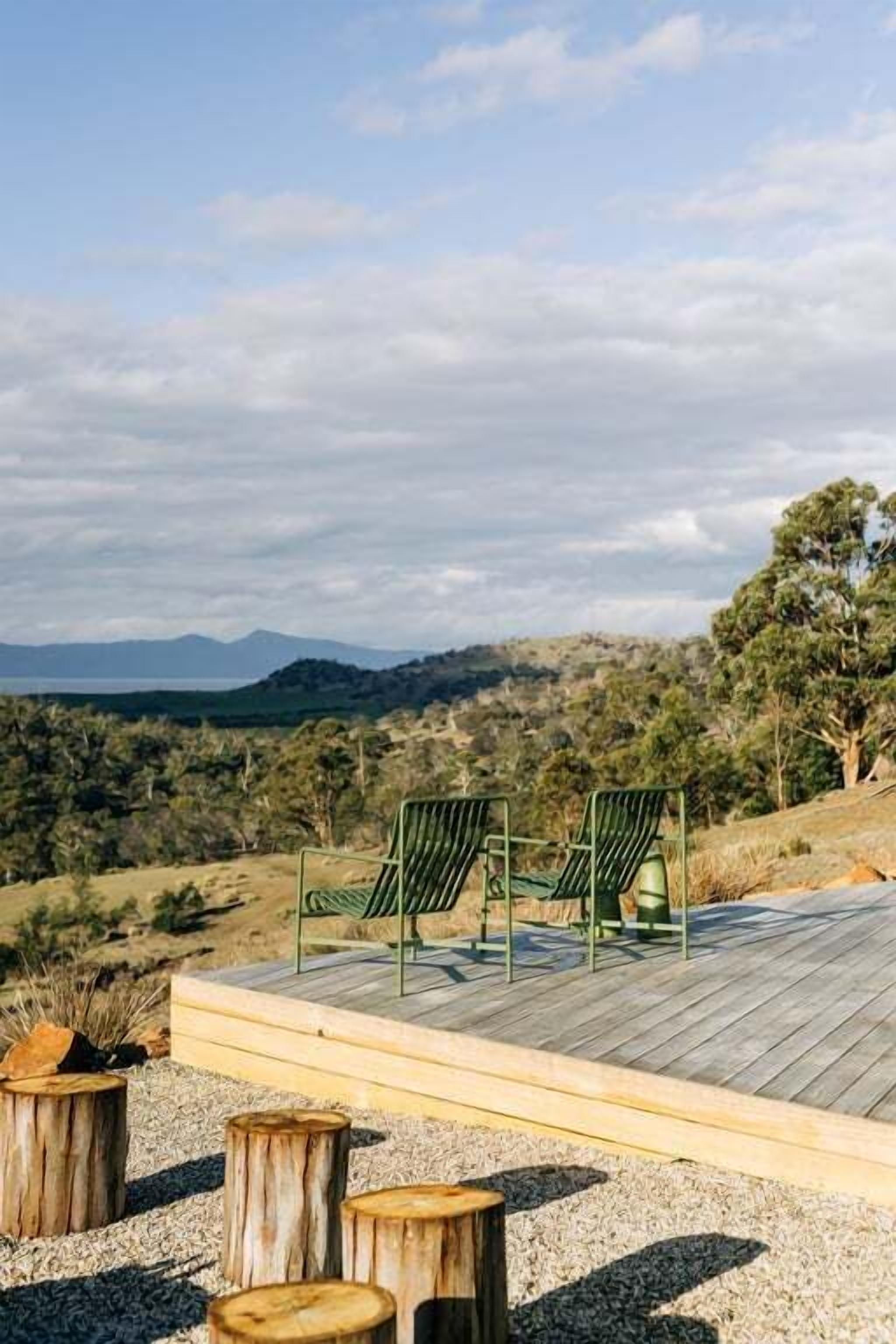 A deck with two chairs and a table, surrounded by wooden stumps, overlooking a hilly landscape.