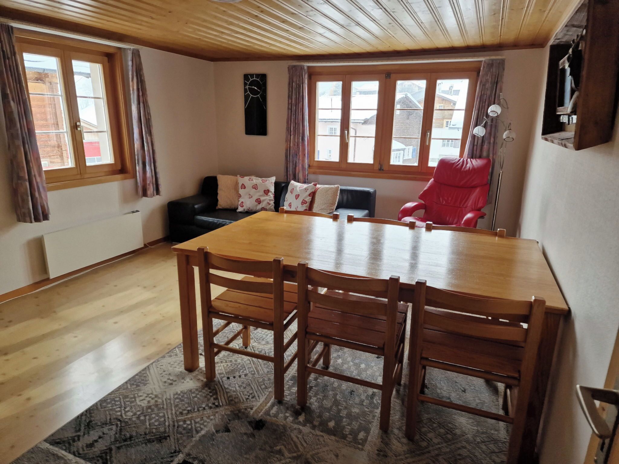 Un'area pranzo con un tavolo e sedie di legno, un divano in pelle nera con cuscini decorati, una sedia rossa e ampie finestre con tende.