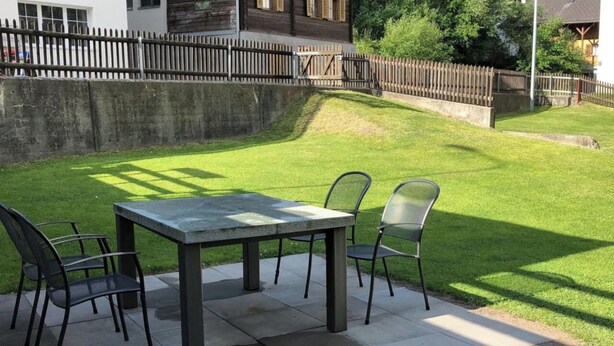 Eine Terrasse im Freien mit Tisch und Stühlen, umgeben von einem gepflegten Rasen und einem Holzzaun.