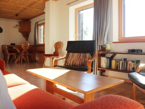 Uma sala de estar acolhedora com uma mesa de madeira, um sofá, uma poltrona com almofada listrada, uma estante de livros e uma janela com cortinas.