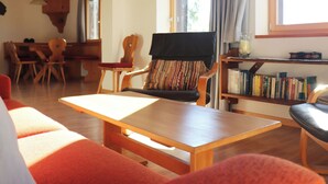 Uma sala de estar acolhedora, com uma mesa de madeira, um sofá e uma poltrona com almofada listrada. Há uma estante de livros e uma janela com cortinas.