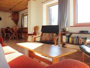 Ein gemütliches Wohnzimmer mit einem Holztisch, einer Couch und einem Sessel mit gestreifter Sitzkissen. Ein Bücherregal mit Büchern und ein Fenster mit Vorhängen sind ebenfalls vorhanden.