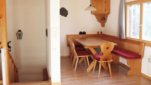 Uma sala de jantar com piso de madeira, mesa e bancos, uma prateleira na parede e uma janela com cortinas.
