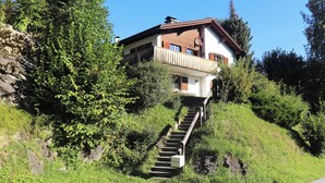 Uma casa de dois andares com varanda, cercada por vegetação e um muro de pedra.