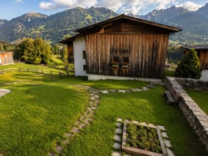 A rustic wooden building with a stone pathway leading to it, surrounded by lush green grass and a mountainous backdrop.