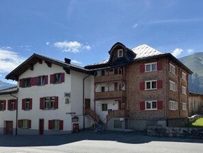 A multi-story building with red shutters, a balcony, and a mountain backdrop.