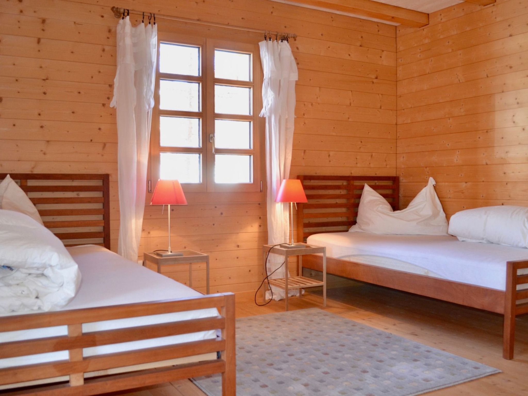 A wooden cabin room with two beds, a window with curtains, and two bedside lamps.