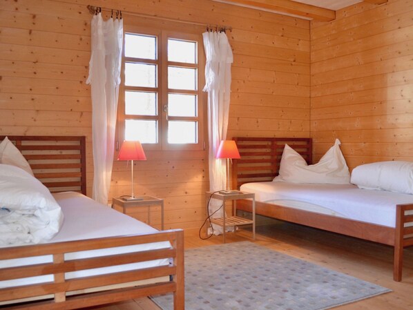 A wooden cabin room with two beds, a window with curtains, and a small table with a lamp.
