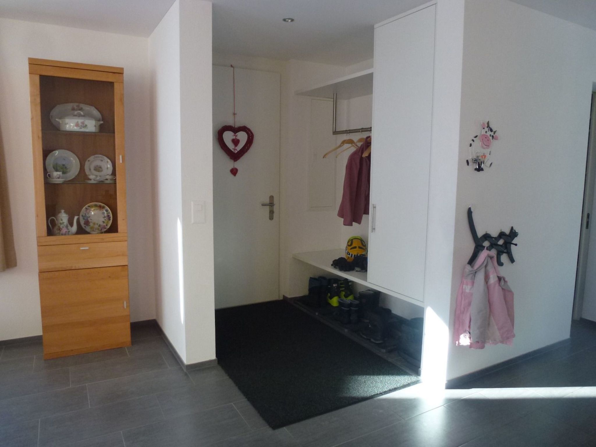 A room with a wooden cabinet containing dishes, a heart-shaped decoration, and a shoe rack with shoes and bags.