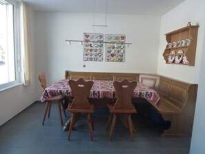 A dining area with a wooden table and chairs, a bench, and a wall-mounted shelf with cups and plates. - Denter Uauls Jucker, Vella, (Vella), 44012B by Interhome (Vella)