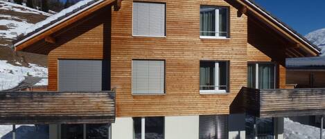 A modern wooden house with a sloped roof, multiple windows, and a covered outdoor area with a table and chairs.