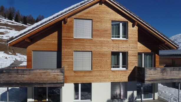 A multi-story wooden house with a sloped roof, large windows, and a covered outdoor area with a table and chairs. - Denter Uauls Jucker, Vella, (Vella), 44012B by Interhome (Vella)