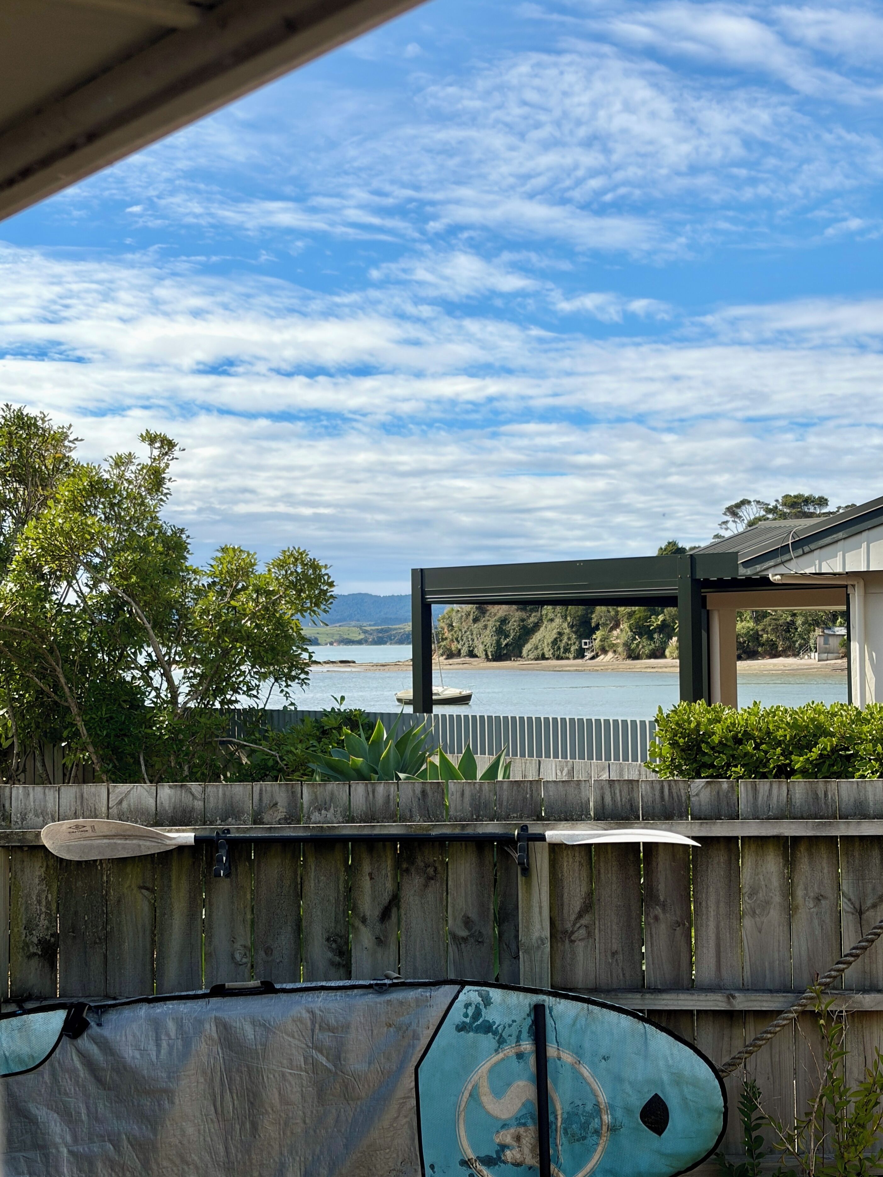 A view of a wooden fence with a paddleboard leaning against it, a wooden deck, and a body of water with a boat in the distance.