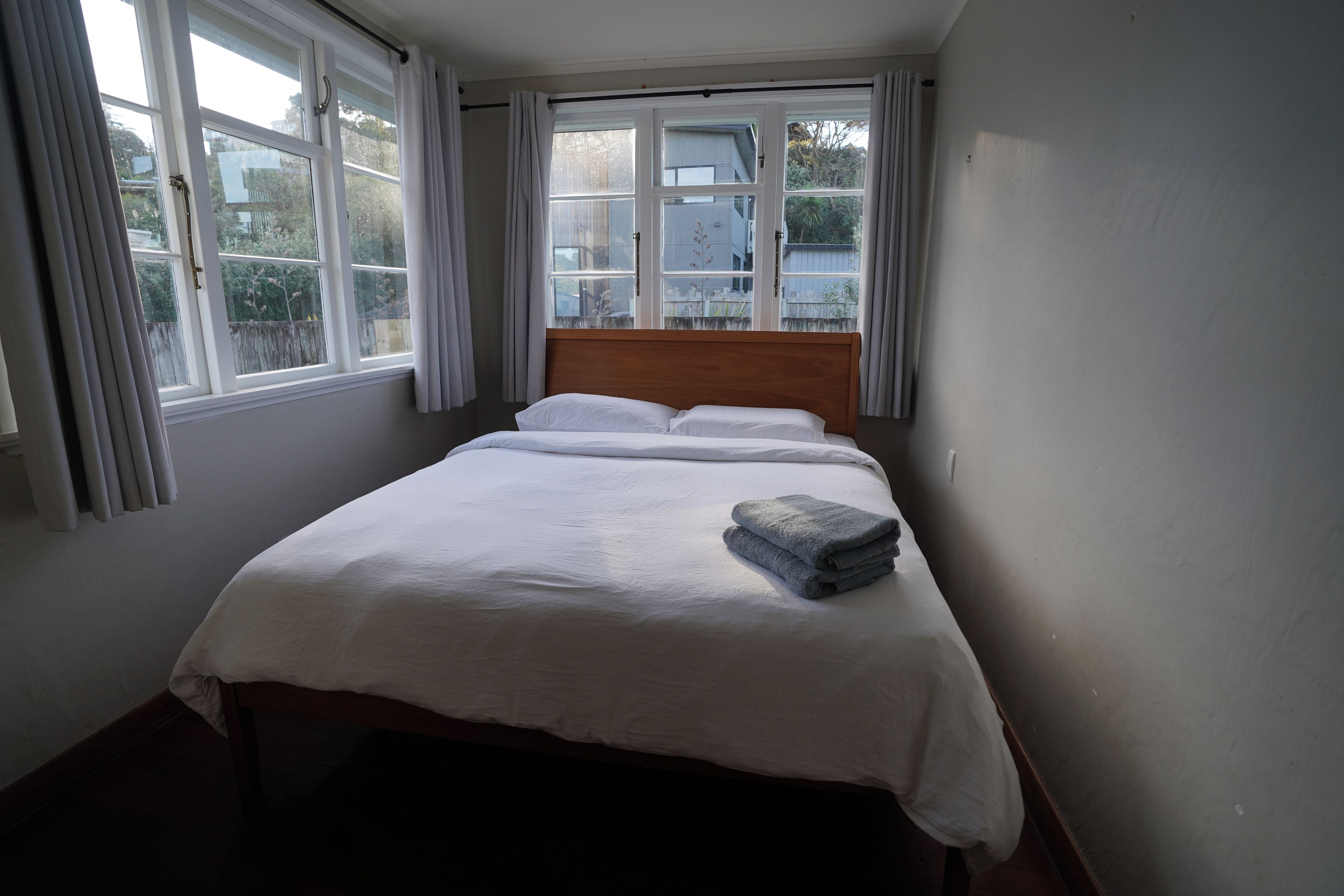 A single bed with white bedding and a folded towel, a wooden headboard, and a window with curtains.
