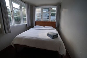 A single bed with white bedding and a folded towel, a wooden headboard, and a window with curtains. - Lorenzen Bay Bungalow by The Sea (Raglan)