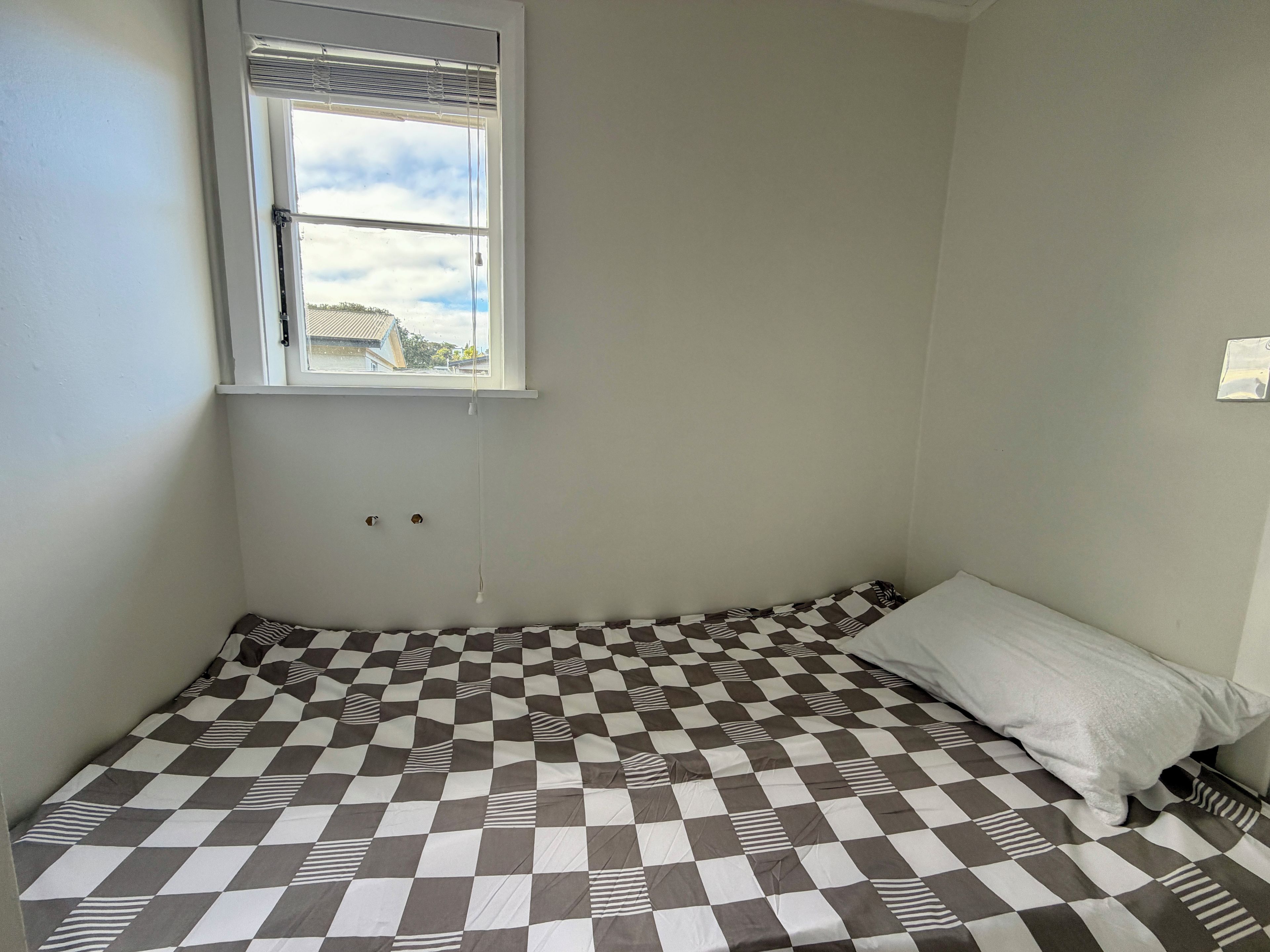 A small, single-bed room with a checkered bedspread, a window with blinds, and a wall-mounted light switch.