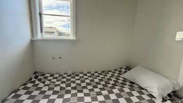 Ein kleines Zimmer mit einem Einzelbett, einer Bettdecke mit Schachmuster, einem Fenster mit Jalousien und einem an der Wand montierten Lichtschalter.