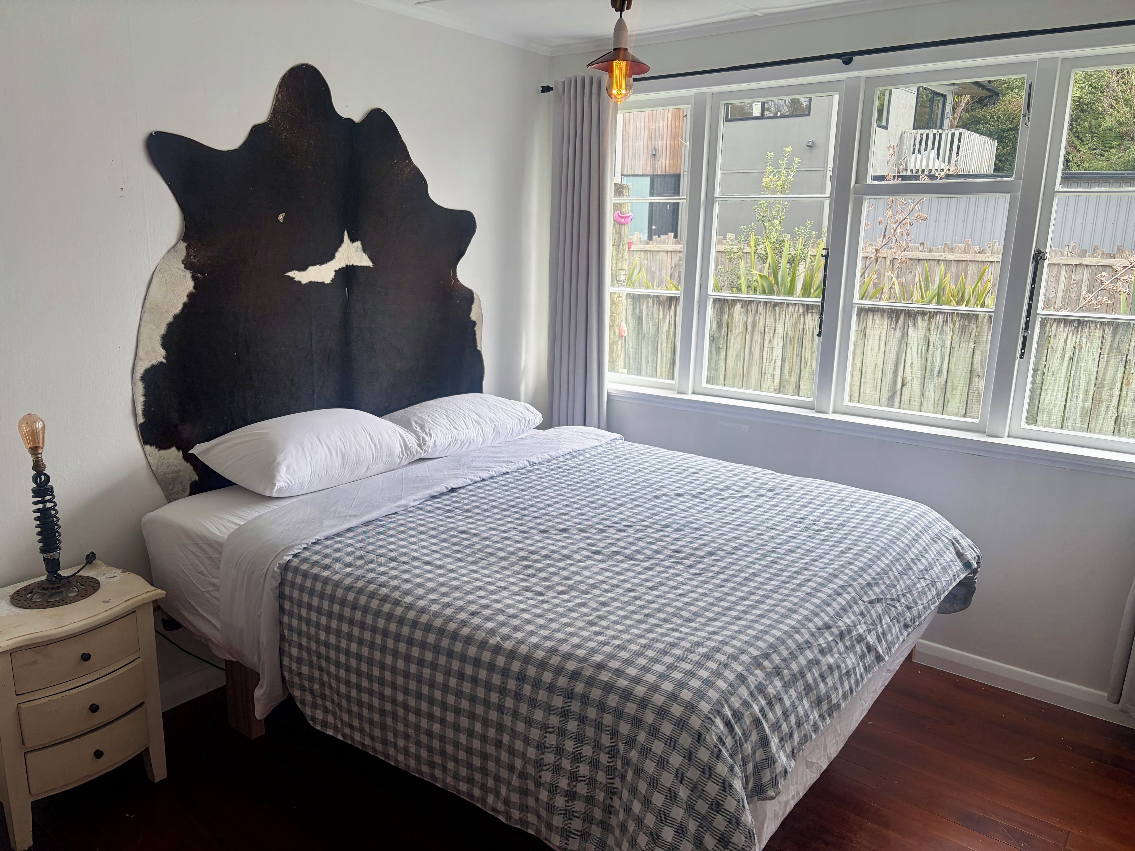 A bedroom with a bed, a nightstand, a wall-mounted headboard, and a window with a view of the outside.