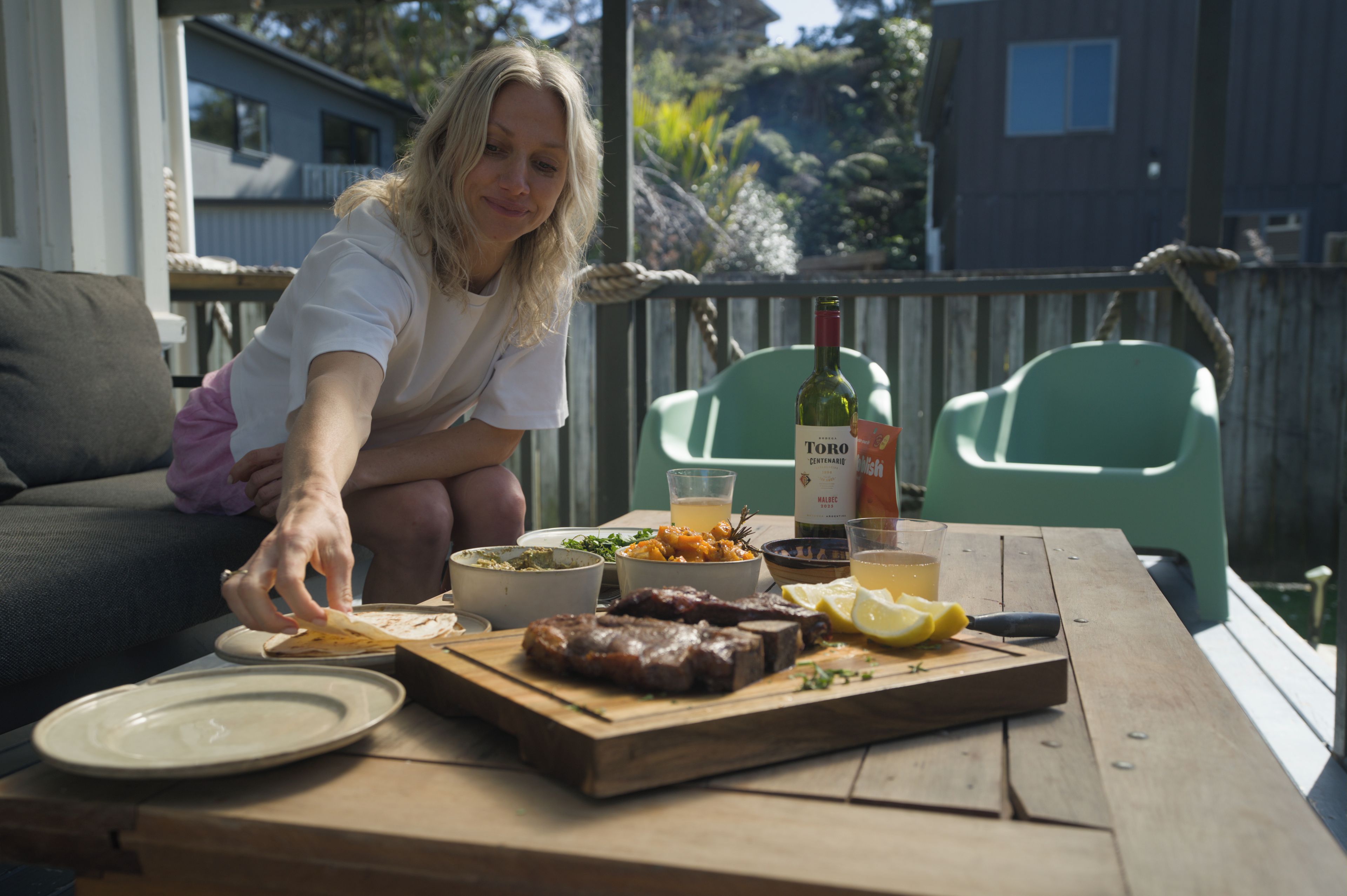 A person serving food on a wooden table outdoors with a bottle of wine, bowls, and plates.
