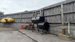 A barbecue grill with a hose, a yellow boat, and a pile of firewood against a wooden fence. - Lorenzen Bay Bungalow by The Sea (Raglan)