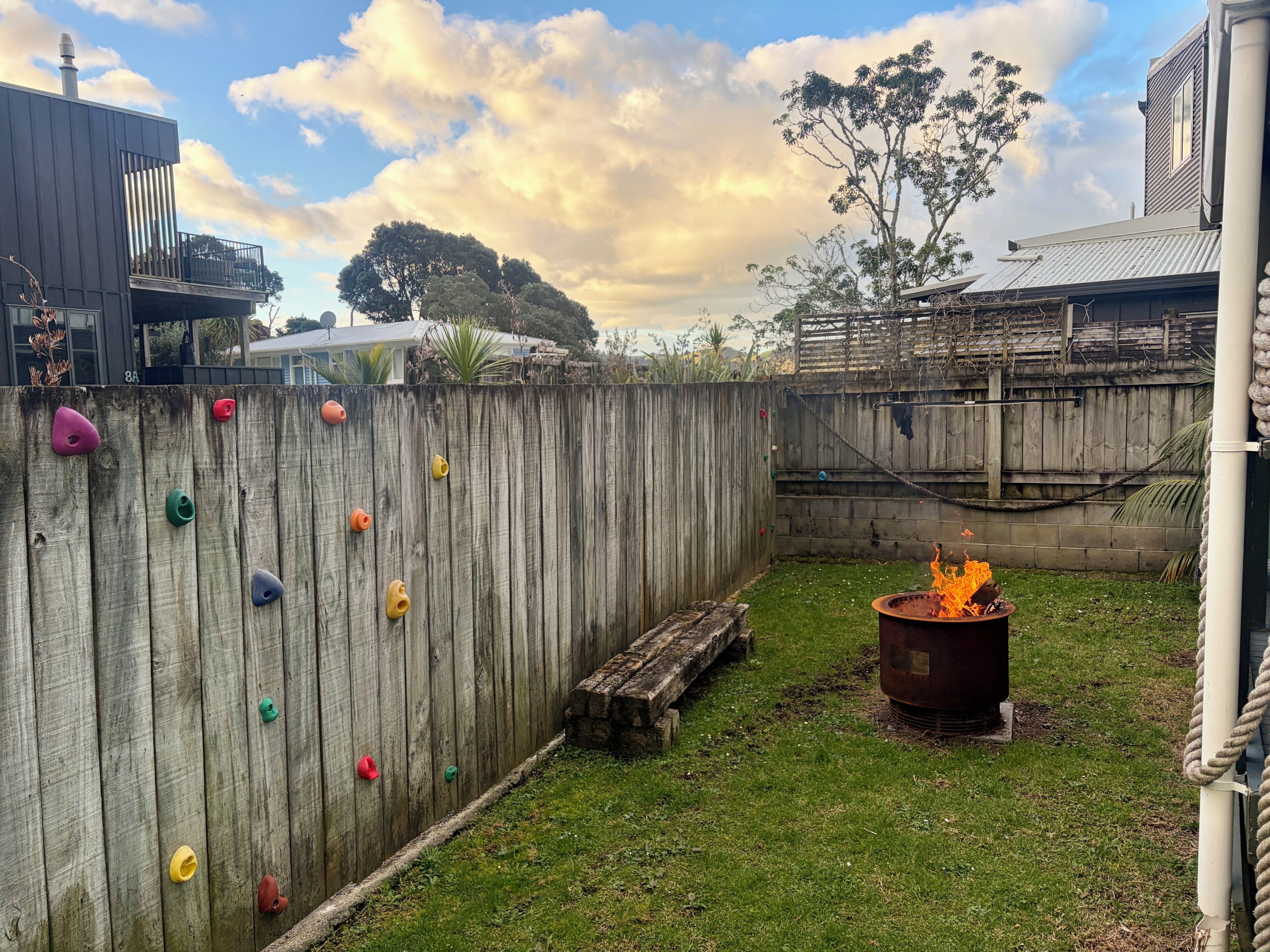Ein Garten mit einer hölzernen Kletterwand, einer Feuerstelle und einer Rasenfläche.
