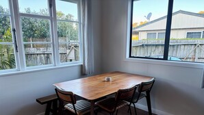 A dining area with a wooden table and four chairs, a bench, and a window with a view of a backyard and a building. - Lorenzen Bay Bungalow by The Sea (Raglan)