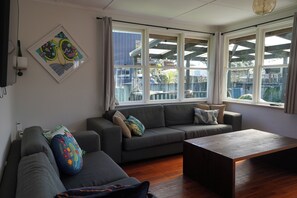 A living room with a grey sofa, a wooden coffee table, and a large window with curtains. - Lorenzen Bay Bungalow by The Sea (Raglan)