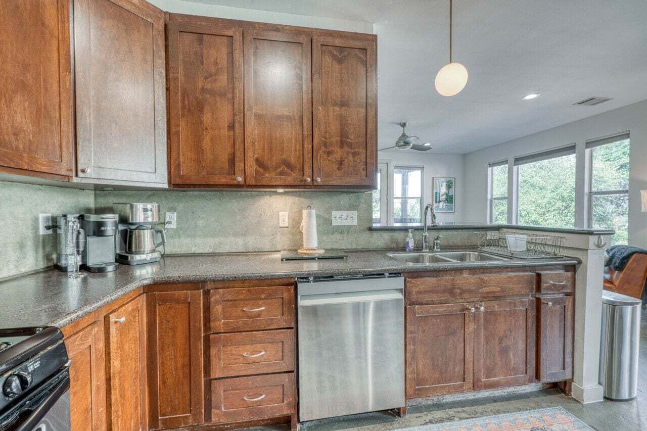 Een keuken met houten kastjes, roestvrijstalen apparatuur, een vaatwasser en een groot raam dat voor natuurlijk licht zorgt.