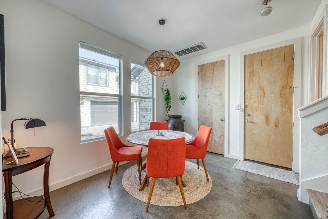 Uma sala de jantar com uma mesa redonda, quatro cadeiras, um lustre e vista para o exterior através de amplas janelas.