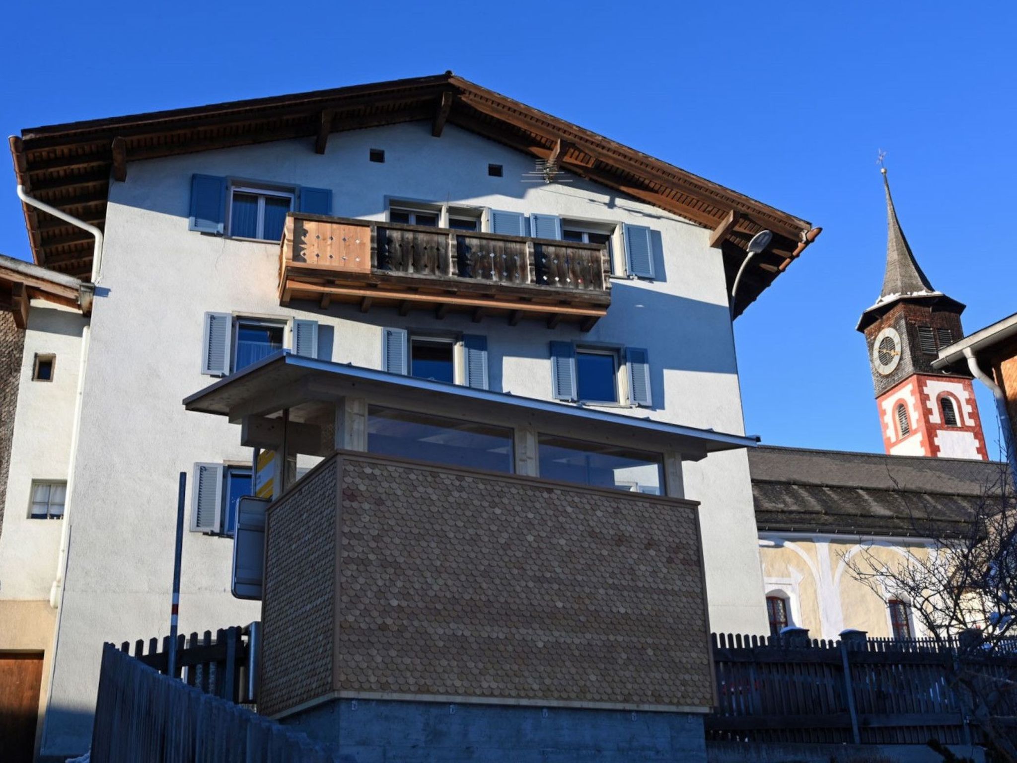 A multi-story building with a balcony, a clock tower in the background, and a clear blue sky.
