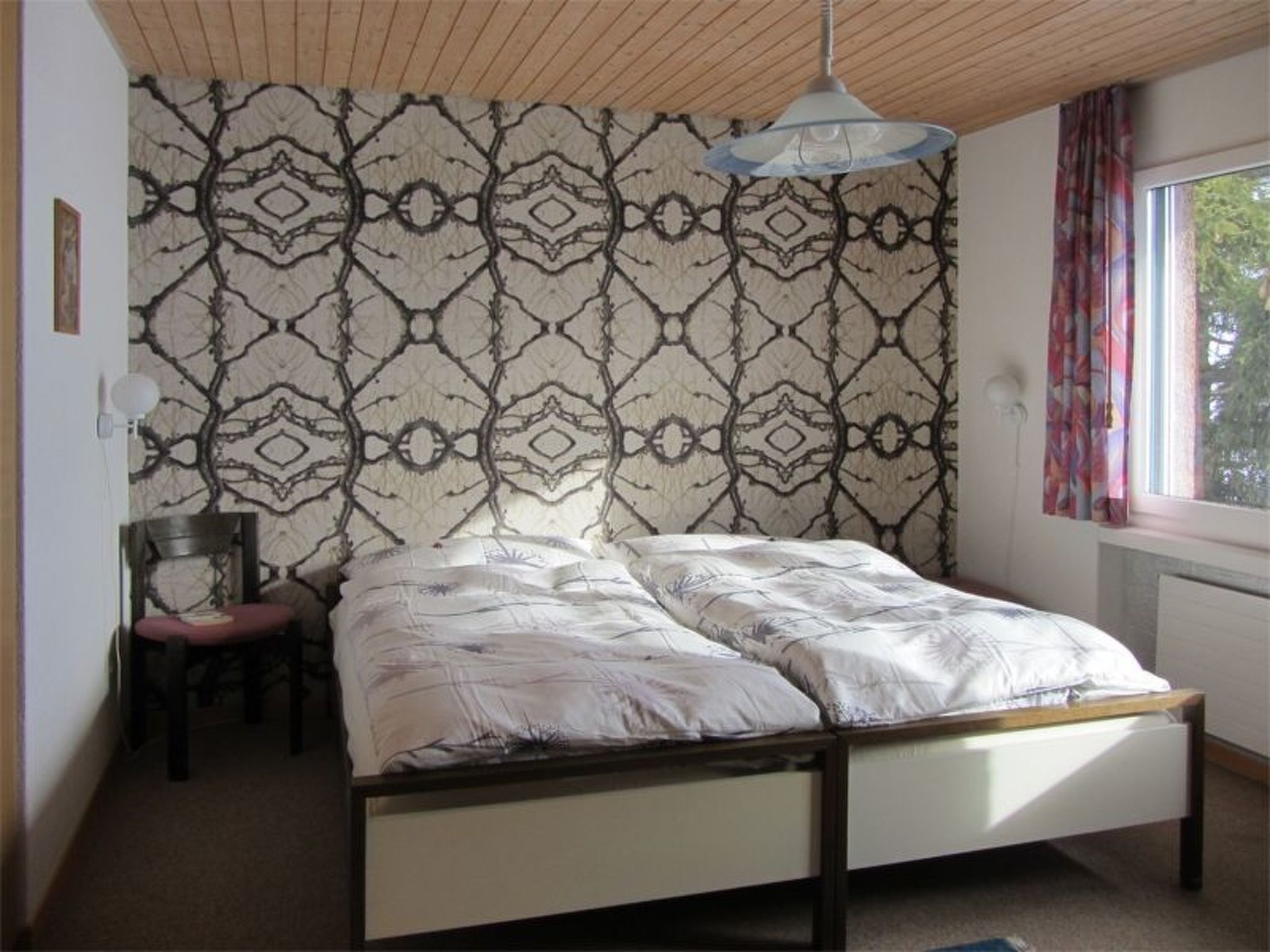 Una cama individual con estructura de madera en una habitación con papel tapiz estampado y una ventana con cortinas.