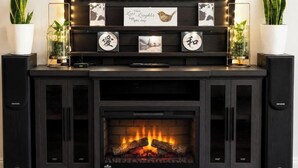 A fireplace with a fire, flanked by two speakers, and a shelf above with framed pictures and a sign. - Your Home Away From Home in Courtice (Clarington)