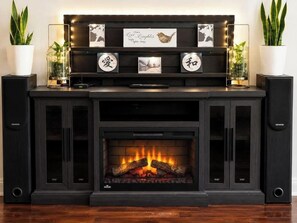 A fireplace with a fire, flanked by two speakers, and a shelf above with framed pictures and a sign.