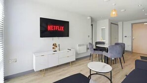 Ein modernes Wohnzimmer mit Flachbildfernseher, einem weißen Schrank, einem Essbereich mit Stühlen und einem kleinen runden Tisch.
