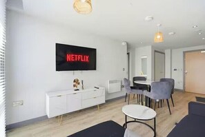 Ein modernes Wohnzimmer mit Flachbildfernseher, einem weißen Schrank, einem Essbereich mit Stühlen und einem kleinen runden Tisch.
