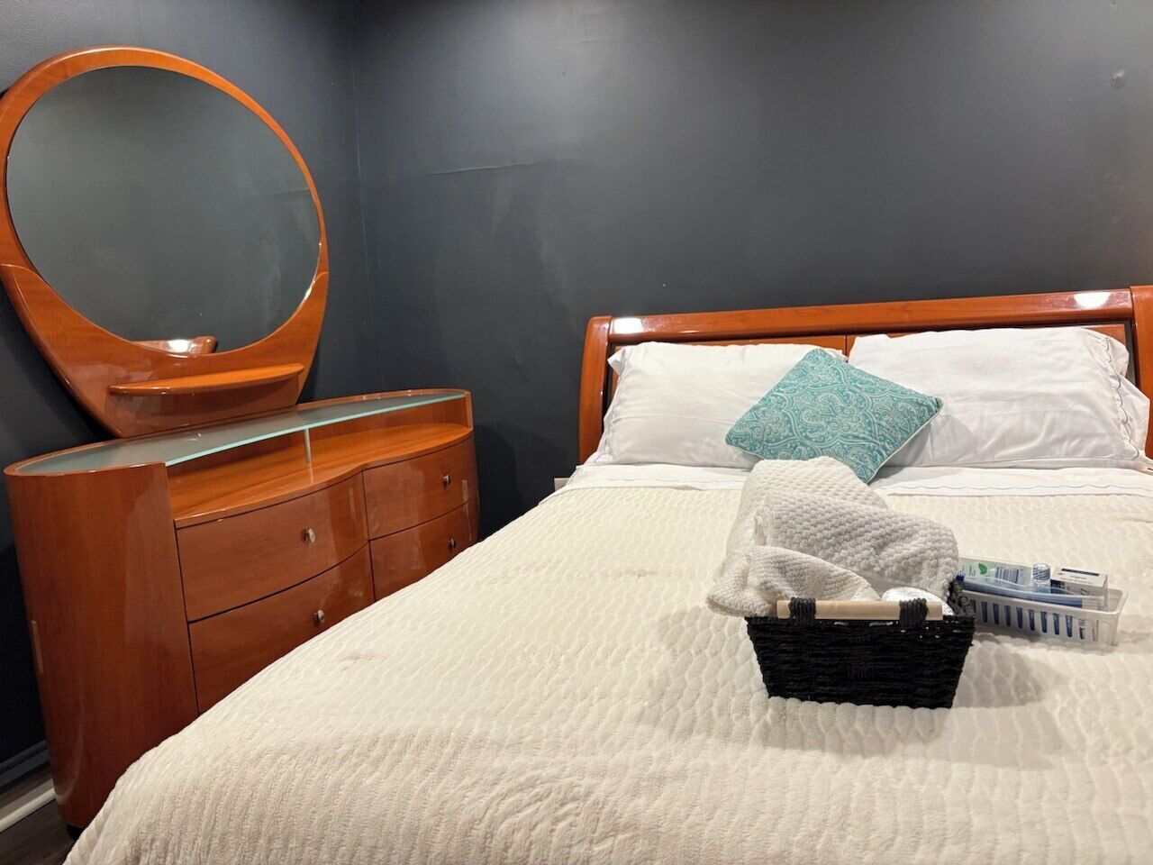 A neatly made bed with a white quilt, a wooden nightstand with drawers, a round mirror, and a bedside table with a basket containing toiletries.