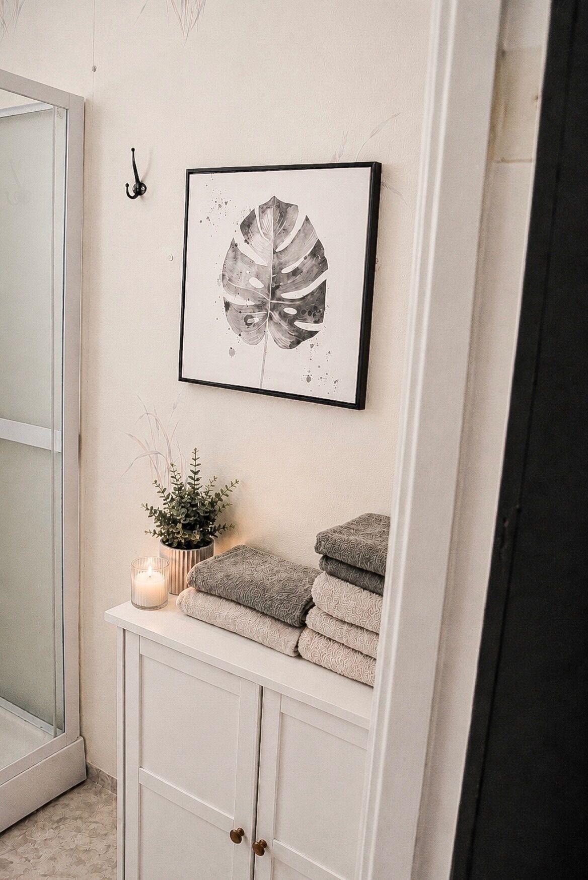 Un baño con una obra de arte de hojas enmarco, una planta en maceta, una vela y toallas dobladas en un mueble.