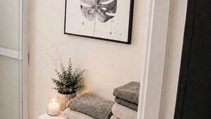 Un bagno con un quadro raffigurante una foglia, una pianta in vaso, una candela e asciugamani riposti ordinatamente in un mobiletto.