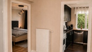 Una camera da letto con un letto e una lampada da comodino, una cucina con elettrodomestici e una finestra, e un corridoio con un termosifone.