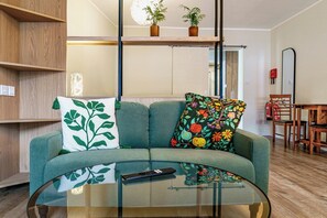 A modern living room with a glass coffee table, a sofa with decorative pillows, wooden shelves, a dining area with a chair, and a fire extinguisher on the wall. - Studio Sleeps 2 with Parking (Belfast)
