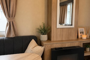 A modern hotel room with a dark-colored sofa, a flat-screen TV, a wooden console table with a potted plant, a framed picture, and a lit candle. - Serene Lodge Retreat in Clacton (Clacton-on-Sea)